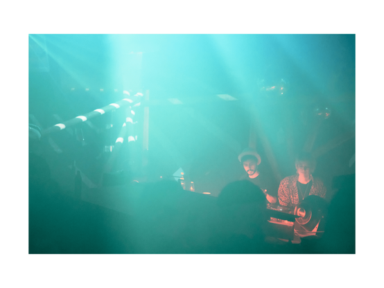 two people djing amid wooden structures with haze and light in the colors mint and orange