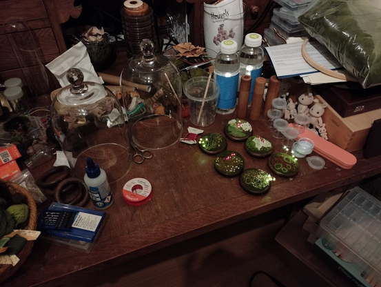 A cluttered wooden workspace filled with various crafting materials and tools. In the center, several small handmade mossy landscapes with embedded LED lights glow softly. Two glass domes sit nearby, along with a bottle of glue, a spool of soldering wire, a pair of scissors, and a set of precision screwdrivers. To the right, a group of Sylvanian Families rabbit figures is gathered next to small plastic containers. A large bag of green wool and embroidery supplies, including a hoop, are stacked in the background. Bottles and wooden cylindrical objects are also present, along with scattered crafting supplies.