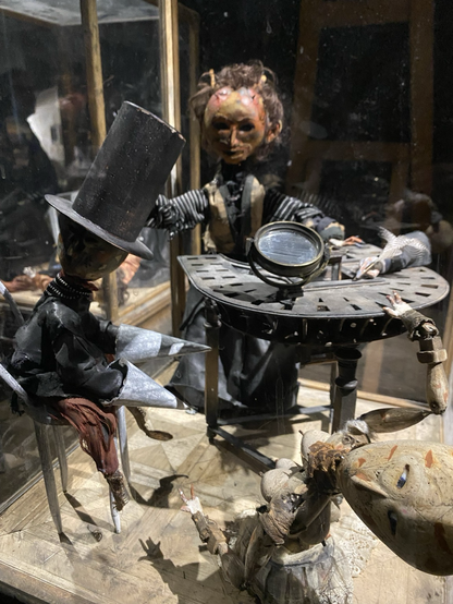 Two dolls sit a table. A third is leaning backwards.