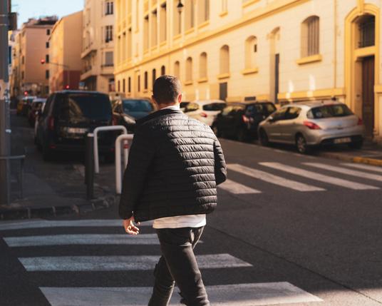 The image shows a person walking on a crosswalk in an urban setting. The individual is wearing a black quilted jacket and appears to be heading towards a street lined with parked cars and buildings. The lighting suggests it might be either morning or late afternoon.