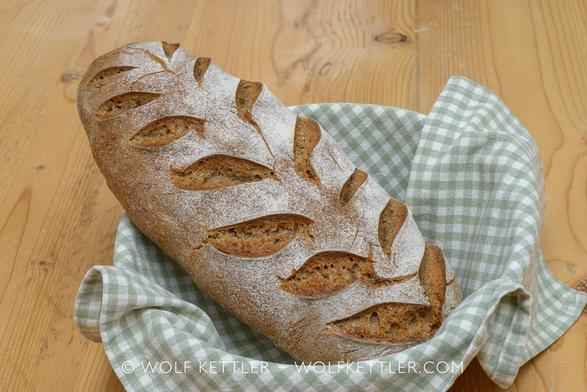A loaf of bread with sheaf-like scoring in a basket. The basket is lined with a green and white checked piece of cloth and the whole arrangement sits on a wooden table top.
