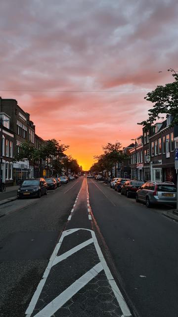 The view down an empty, two lane, black asphalt road with white markings running down the middle and sidewalk parked cars and a mix of various two and three story buildings on both sides. All this under a sky that starts in soft pink with white cloud swirls and gradually turns more orange and then bright yellow as the sun sets at the far off horizon.