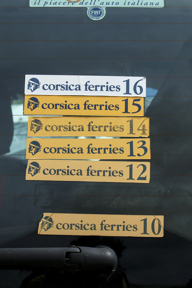 das hochformat zeigt auf der heckscheibe eines autos verschiedene aufkleber der corsica ferries, die von angestellten der fährgesellschaft ungefragt auf die autoscheiben bei der überfahrt nach korsika angebracht werden.
der unterste ist 'corsica ferries 10', der oberste 'corsica ferries 16'. dazwischen sind alle anderen jahresaufkleber zu sehen. nur der aufkleber 'corsica ferries 11' scheint abgefallen oder entfernt worden zu sein. dieser fehlt. 
die aufkleber 10–15 sind mit schwarzer schrift auf gelben grund, der aufkleber 16 ist auf weißem grund.