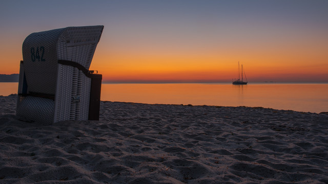 Die Sonne geht über der Ostsee auf, auf dem Sandstrand steht links ein Strandkorb. Im Hintergrund steht ein Segelboot auf dem Wasser. Der Himmel hat die orangeroten Töne die typisch für den Sonnenaufgang sind. 