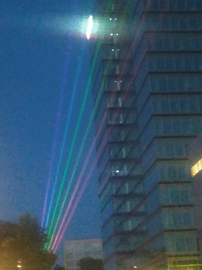 Regenbogenlaserstrahlen fächerförmig vom unteren Bildrand nach oben vor dunkelblauem Himmel. Am rechten Bildrand ein Hochhaus, am unteren Bildrand Bäume und andere Gebäude