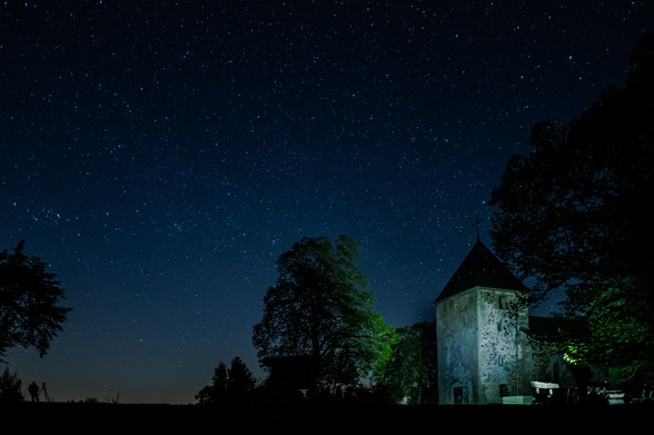 Das Foto zeigt einen gigantischen Sternenhimmel in Wollseifen im Nationalpark Eifel. Da auch umliegende Orte ihre Nachbeleuchtung runter drehen, ist der Park ein Teil der dark sky Community. Ideal zum Sterne fotografieren.
Unten rechts im Bild ist die Kirche St.Rochus zu sehen, sowie ein Bank zum rasten. Ich beleuchtete beides während der Langzeitbelichtung mit einer starken Taschenlampe, so das man die Kirche sehr gut erkennen kann. Normalerweise wäre dort alles dunkel. 
Weiter sind ein paar Bäume am unteren Bildrand zu sehen, sowie links unten mein Fotofreund Dietmar der eine Kamera mit Stativ vor sich stehen hat. 