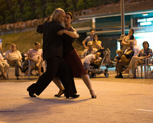 Ein Mann und eine Frau tanzen Tango, um sie herum sitzen Zuschauer und schauen den beiden zu. Die Szene spielt in Italien an einem Sommerabend.