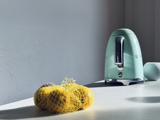 A light green kettle is positioned on a white countertop next to a mesh bag containing three lemons. The scene is softly illuminated with a shadow cast across the surface.