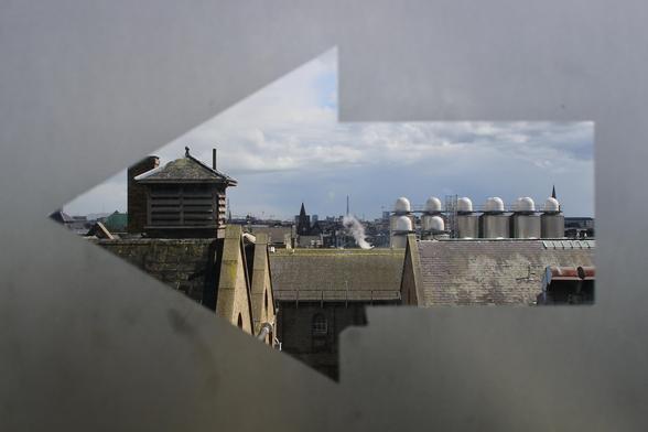 das bild im querformat zeigt durch eine pfeilförmige aussparung einer milchglasscheibe den blick auf die dächer einer stadtlandschaft.
der pfeil zeigt nach links. die milchglasscheibe ist undurchsichtig. die aussparung erlaubt den blick nach draußen. im vordergrund sind die dächer von gebäuden mit ziegeldächern zu sehen. dahinter sind industrieanlagen erkennbar. rauch steigt auf. je weiter der blick in die tiefe geht, desto mehr städtische gebäude und dächer sind sichtbar. der himmel ist bewölkt.