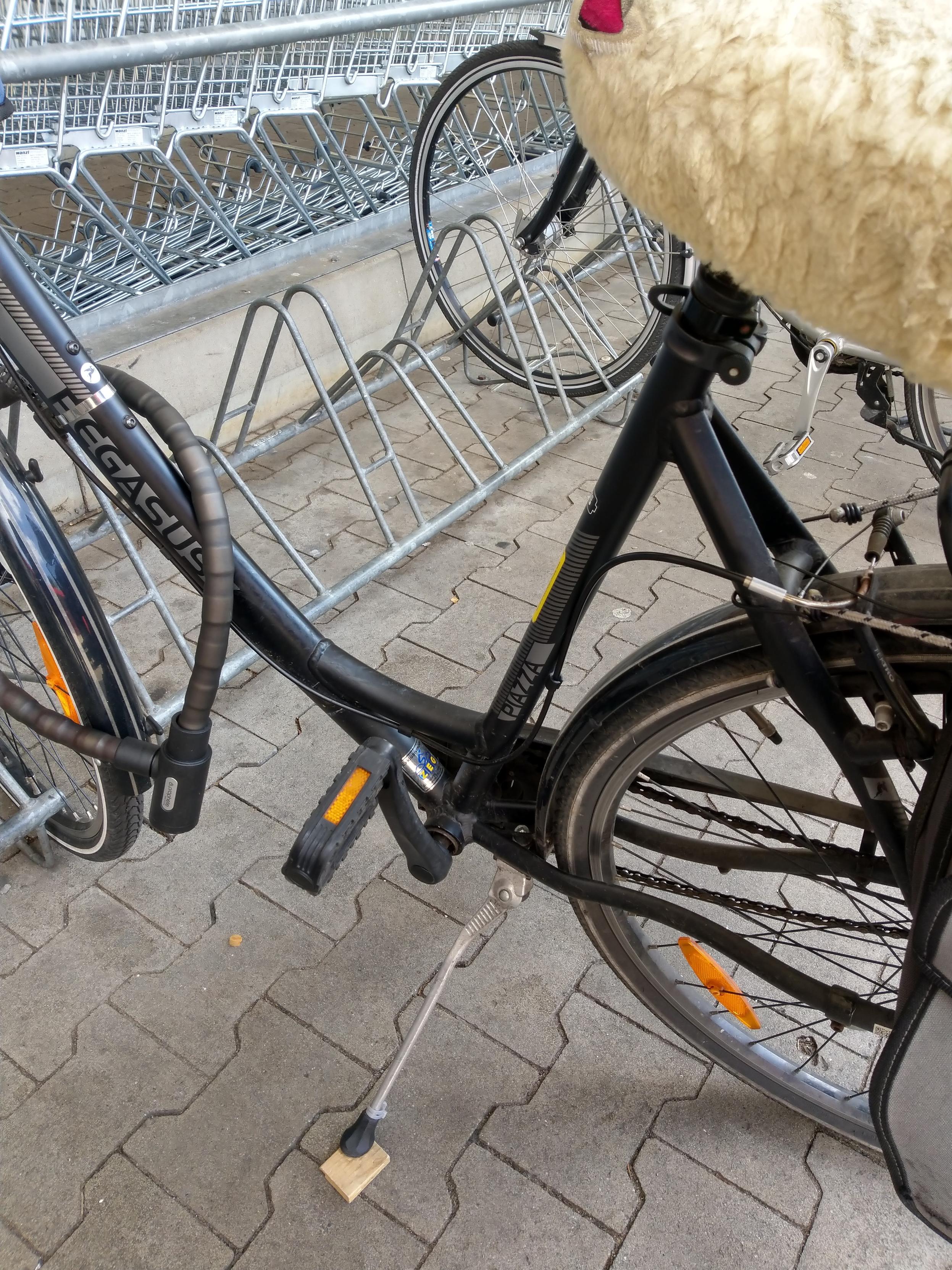 Teilansicht eines Fahrrads mit Tiefeinstieg an den Fahrradständern vor einem Supermarkt.
Weil der Seitenständer zu kurz ist, hat der Eigentümer, die Eigentümerin ein Holzplättchen dabei und legt es unter den Seitenständer.
🤷🏻‍♂️
Man muss sich nur zu helfen wissen.
Oben rechts ragt der Sattel mit einem hellen Fellüberzug ins Bild.