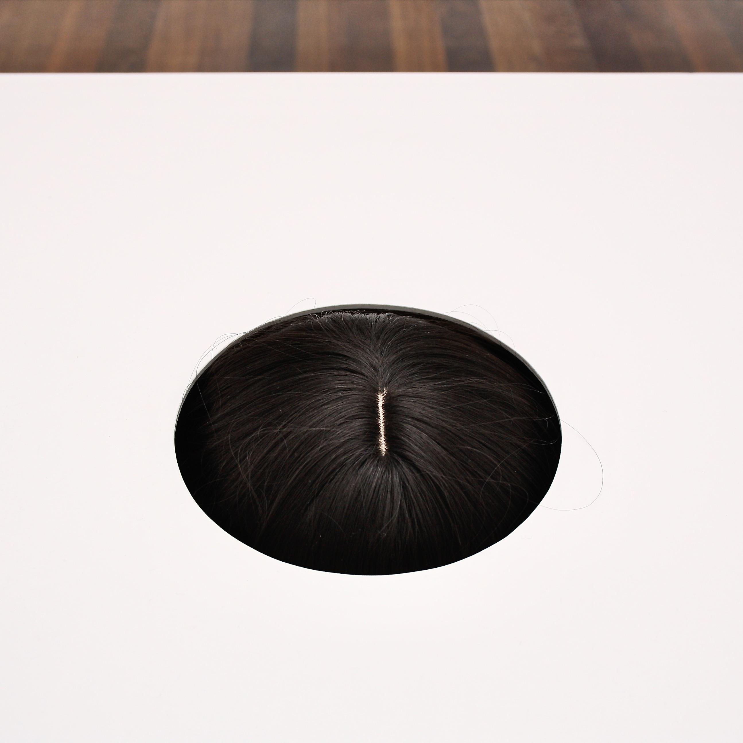 das quadratische bild zeigt den oberen teil einer dunkelbraunen haarpracht, die durch ein kreisförmiges loch in einer weißen oberfläche sichtbar ist. am oberen bildrand ist der fußboden hinter der weißen platte zu sehen. es ist ein schmaler streifen braunen parketts. 
das bild ist minimalistisch und der fokus liegt ganz auf dem kontrast zwischen der dunklen haarpracht in dem runden loch und der hellen, weißen oberfläche.
es wirkt ruhig und etwas geheimnisvoll.
