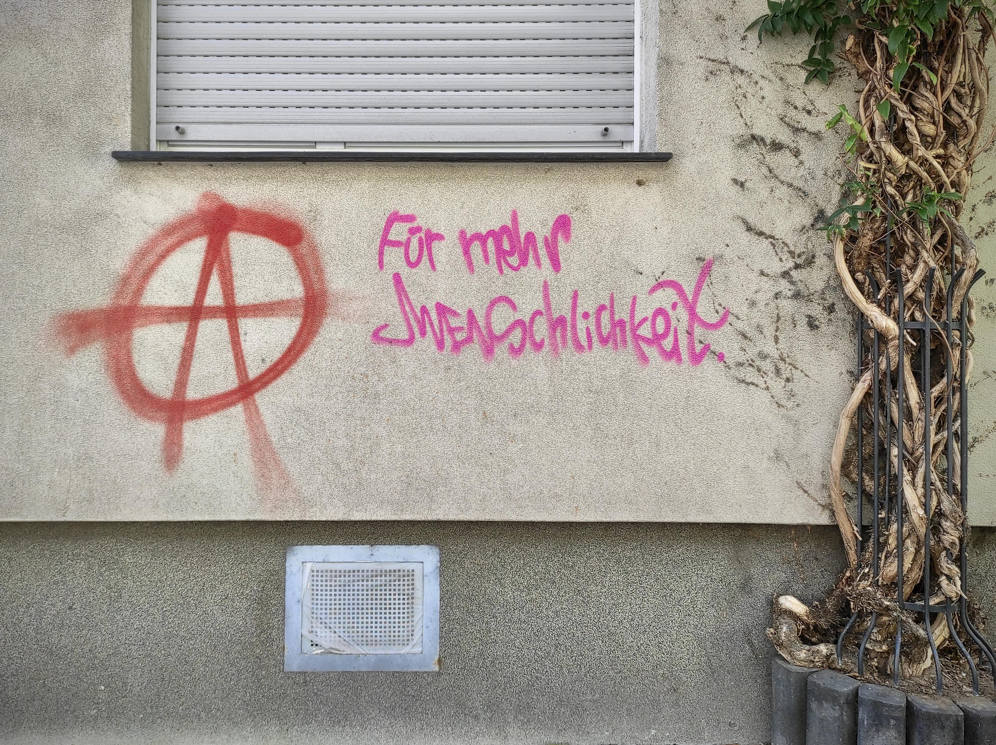 das bild zeigt ein stück einer hauswand aus hellem verputz. oben ist ein fenster angeschnitten, dessen rolläden heruntergelassen sind. darunter ist ein anarchiezeichen in rot, also ein kreis mit einem großen "a" darin, und daneben ist in pink der text "für mehr menschlichkeit" gesprüht. rechts neben dem spruch wächst eine pflanze an einem gitter. unter dem spruch befindet sich ein lüftungsgitter.
das gesprühte in rot und pink hebt sich farblich vom rest deutlich hervor und bildet das zentrale element des bildes. das bild zeigt ein stück einer hauswand aus hellem verputz. oben ist ein fenster angeschnitten, dessen rolläden heruntergelassen sind. darunter ist ein anarchiezeichen in rot, also ein kreis mit einem großen "a" darin, und daneben ist in pink der text "für mehr menschlichkeit" gesprüht. rechts neben dem spruch wächst eine pflanze an einem gitter. unter dem spruch befindet sich ein lüftungsgitter.
das gesprühte in rot und pink hebt sich farblich vom rest deutlich hervor und bildet das zentrale element des bildes.