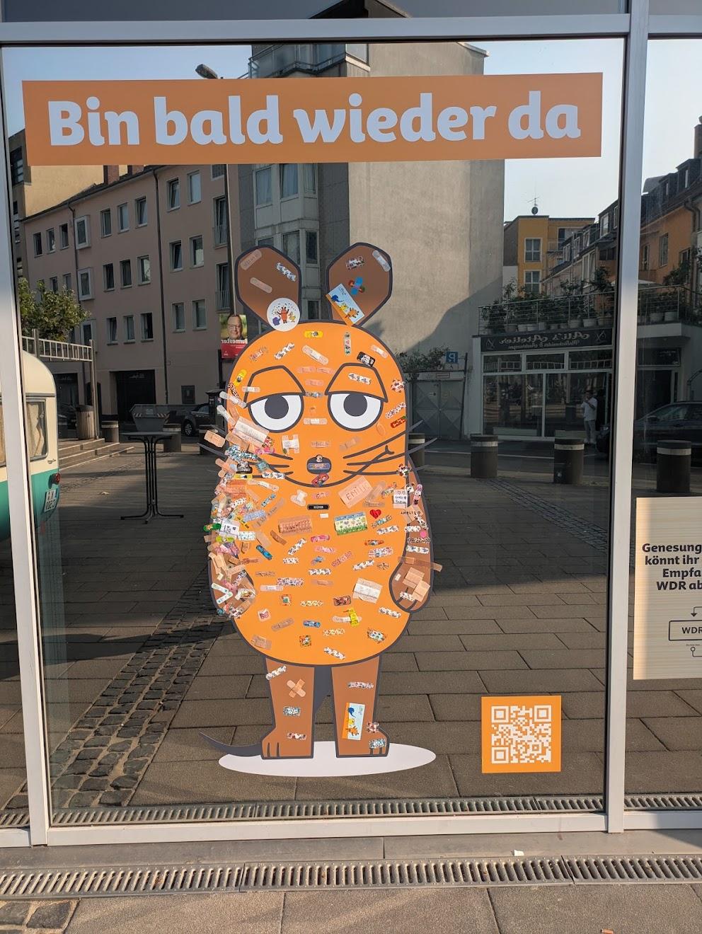 Eine Maus in Orange, aufgeklebt auf ein Schaufenster, mit Schriftzug darüber "Bin bald wieder da". Auf den Maus-Aufkleber sind Dutzende Pflaster aufgeklebt