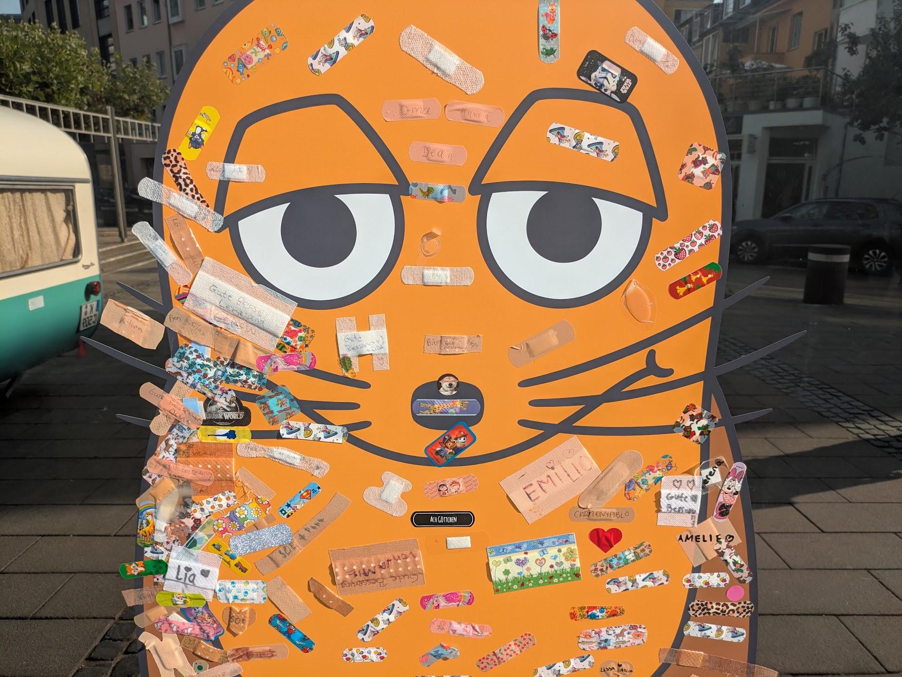 Eine Maus in Orange, aufgeklebt auf ein Schaufenster". Auf den Maus-Aufkleber sind Dutzende Pflaster aufgeklebt