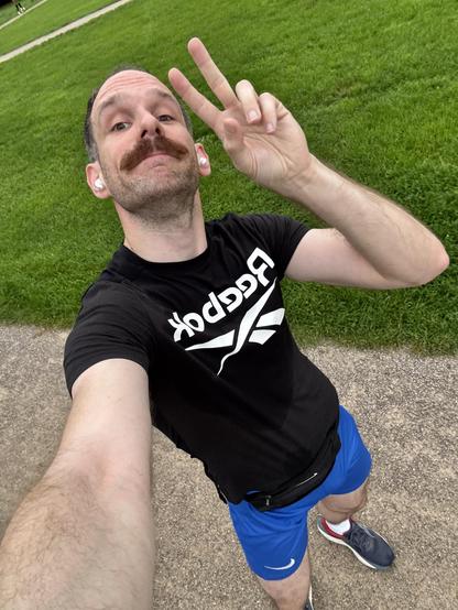 Mann mit Schnurrbart macht Peace-Zeichen, trägt schwarze Reebok-T-Shirt und blaue Shorts, steht auf einem Gehweg.
