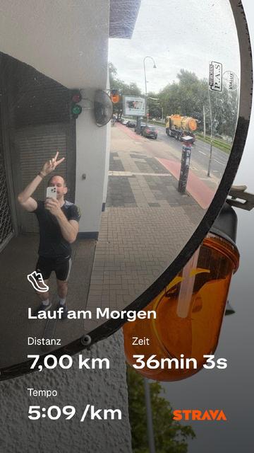 Ein Mann in Laufkleidung macht ein Selfie vor einem großen Verkehrsspiegel an einer Straßenecke. Er zeigt das Victory-Zeichen und trägt ein dunkles Laufshirt. Im Spiegel spiegelt sich die Straßenszene mit Radweg, Gehweg und Bäumen wider. Über das Bild ist eine Strava-Laufstatistik gelegt, die zeigt: "Lauf am Morgen", Distanz 7,00 km, Zeit 36min 3s, Tempo 5:09/km. Das Strava-Logo ist in der unteren rechten Ecke sichtbar.
