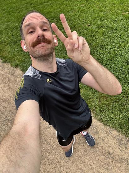Ein Mann mit Schnurrbart und kurzen Haaren liegt auf einer grünen Rasenfläche und macht ein Selfie. Er trägt ein dunkelblaues Sportshirt mit gelben Akzenten und zeigt mit seiner rechten Hand das Victory-Zeichen (Peace-Zeichen) in die Kamera. Im Hintergrund ist gepflegter Rasen zu sehen, und am Bildrand ist ein Gehweg erkennbar.