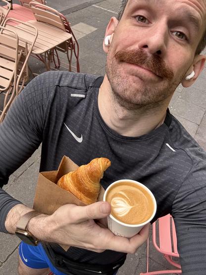 Mann mit croissant und Kaffee auf Straßen café, trägt AirPods und sportliche Kleidung.
