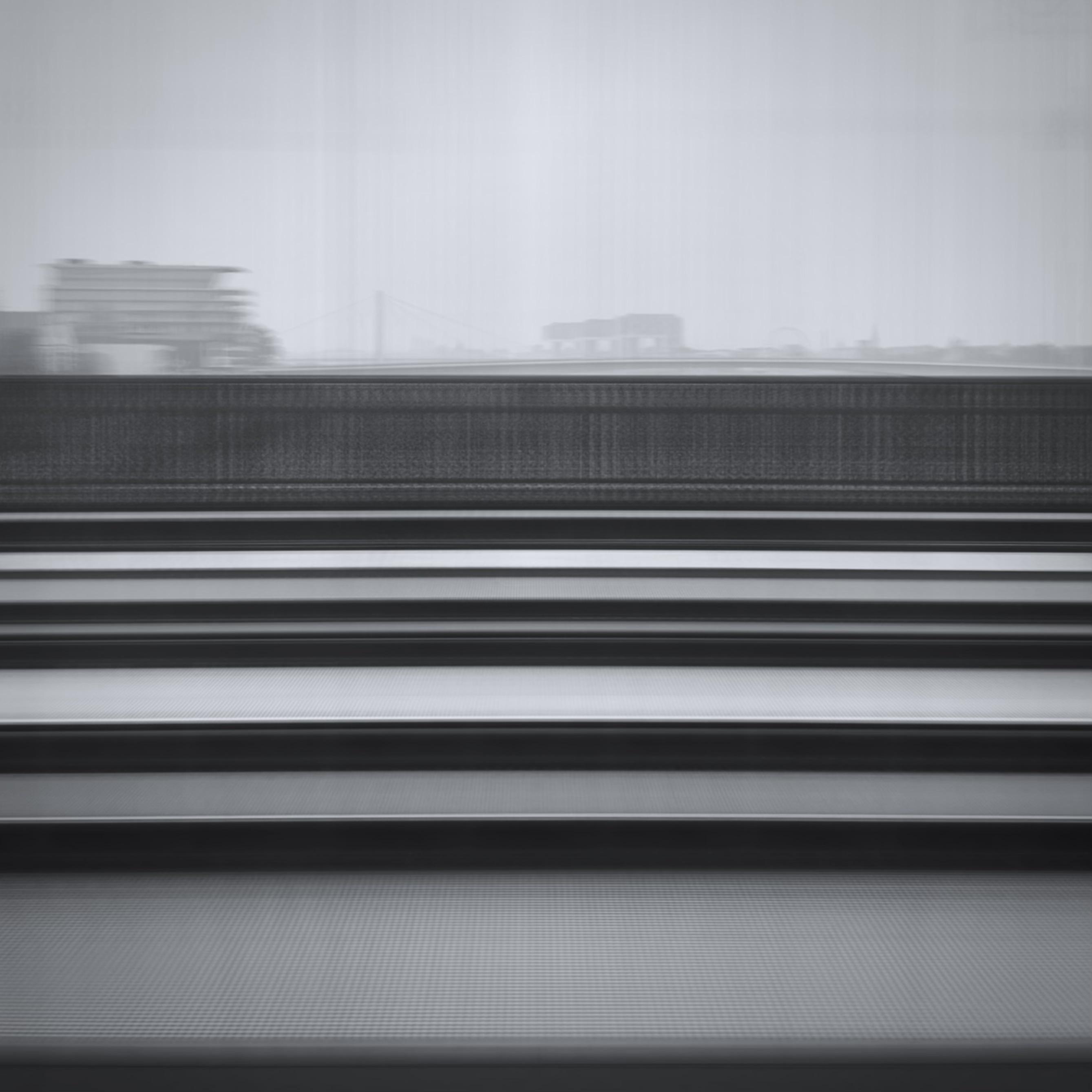 das bild zeigt eine unscharfe, monochrome ansicht aus einem zug heraus. im vordergrund sind horizontale linien zu sehen, die durch gleise und geländer erzeugt werden. im hintergrund sind verschwommene gebäude und eine brücke erkennbar. die kranhäuser in köln sind erkennbar.