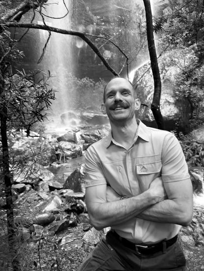 A man with a mustache smiling with arms crossed, standing in front of a waterfall and trees in a natural outdoor setting.