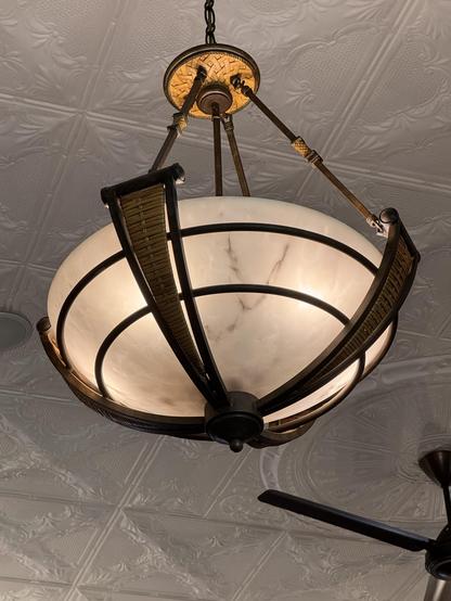 A decorative ceiling light fixture with a frosted glass shade and a bronze metal frame, hanging from a chain on a patterned ceiling.