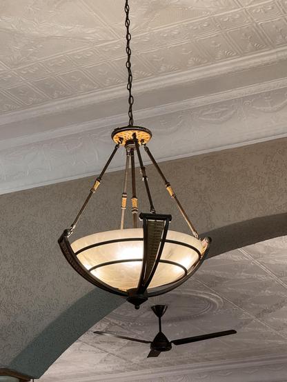 An ornate ceiling light fixture with a frosted glass bowl and bronze accents hangs from a chain, with a ceiling fan visible below.