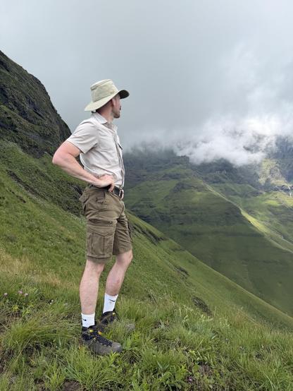 A man dressed in outdoor hiking gear, standing on a grassy hillside with green mountains and cloudy sky in the background.