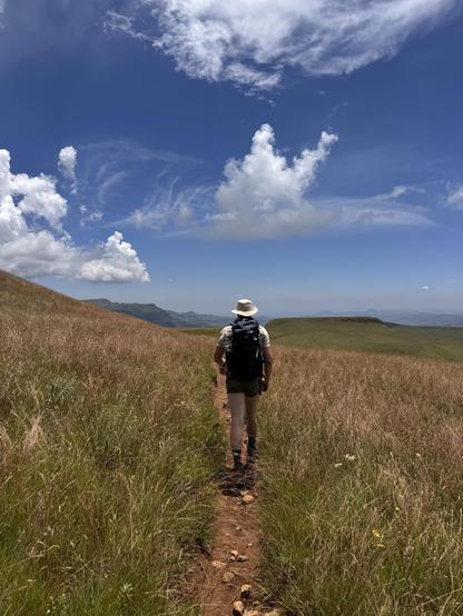 A person with a backpack and hat walking along a narrow dirt trail through a grassy landscape under a partly cloudy blue sky.