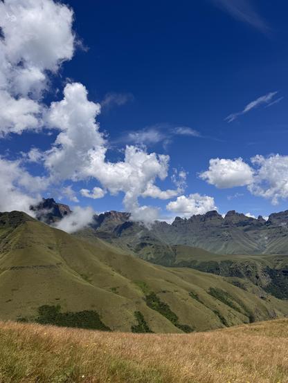 A scenic view of rolling green hills with mountain peaks in the background under a bright blue sky with white, fluffy clouds.