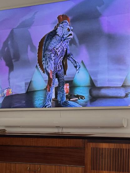 A scene from the 1966 science fiction film "Destination Inner Space" displayed on a television screen, showing an alien creature costume with a textured, scale-like surface in shades of blue and purple. The humanoid creature stands in what appears to be an underwater laboratory set with cone-shaped structures visible in the background. The scene is lit with atmospheric purple and blue lighting, creating an eerie underwater ambiance. The television sits on a wooden media console.