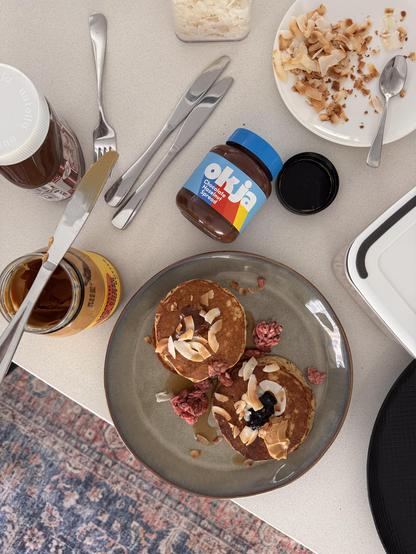 Pfannkuchen mit Schokolade, Kokosnüssen und Beeren auf einem Teller, umgeben von Frühstückszutaten und Besteck.