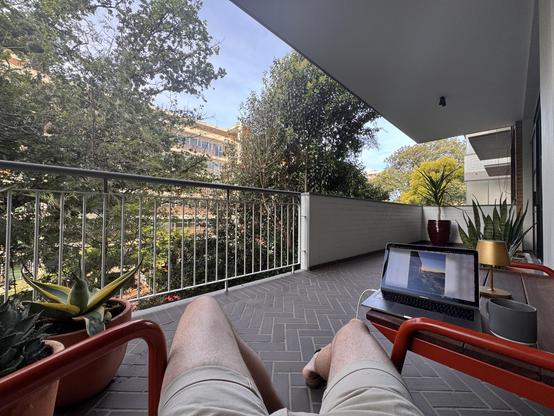 Entspannte Person sitzt auf einem Balkon mit Laptop und Pflanzen, blickt in die Ferne, umgeben von Bäumen und modernen Gebäuden.