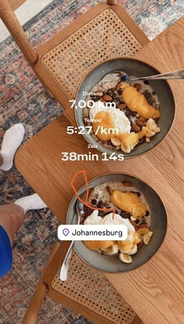 A top-down view of two bowls of oatmeal on a wooden table with cane-back chairs, shot from above. Each bowl is topped with mango purée, sliced banana, almonds, chia seeds, chocolate chips, and a dollop of joghurt. Overlaid text shows running stats: Distance 7.00 km, Pace 5:27 /km, Time 38 min 14 s. A location tag reads "Johannesburg". The feet of a person in white socks are visible at the edge of the frame.