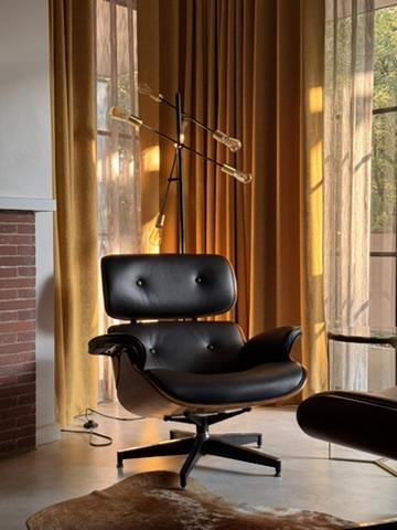 A classic Eames Lounge Chair in black leather with a walnut shell sits on a cowhide rug in a warmly lit living room. Behind it, floor-to-ceiling golden-ochre curtains filter soft afternoon sunlight. A mid-century modern floor lamp with four exposed Edison bulbs on adjustable arms rises behind the chair. A brick fireplace is partially visible on the left, and a side table peeks in from the right.