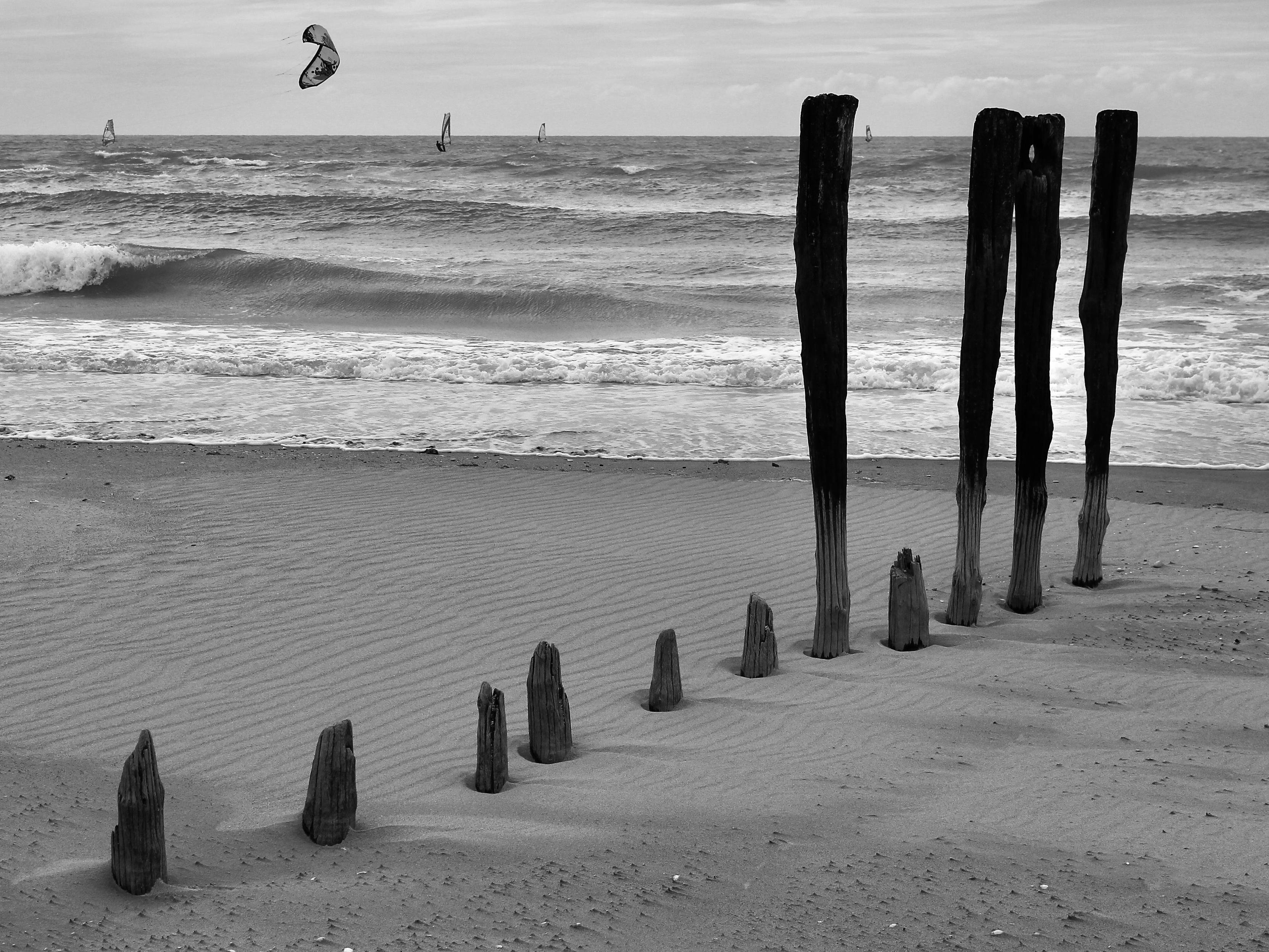 das bild zeigt eine schwarz-weiß-aufnahme eines strandes mit einer reihe von holzpfählen, die aus dem sand ragen.
im sand sind mehrere verwitterte holzpfosten in einer linie angeordnet, die schräg zum wasser führt. die vorderen pfosten sind kurz und abgebrochen, während sie im hintergrund deutlich höher werden und bis über den hoch stehenden horizont ragen.
der sandstrand weist feine wellenmuster auf. dahinter bricht sich die brandung des meeres in schaumigen wellen.
auf dem wasser sind kleine segel von windsurfern zu erkennen. am bewölkten himmel schwebt ein dunkler kite-schirm.