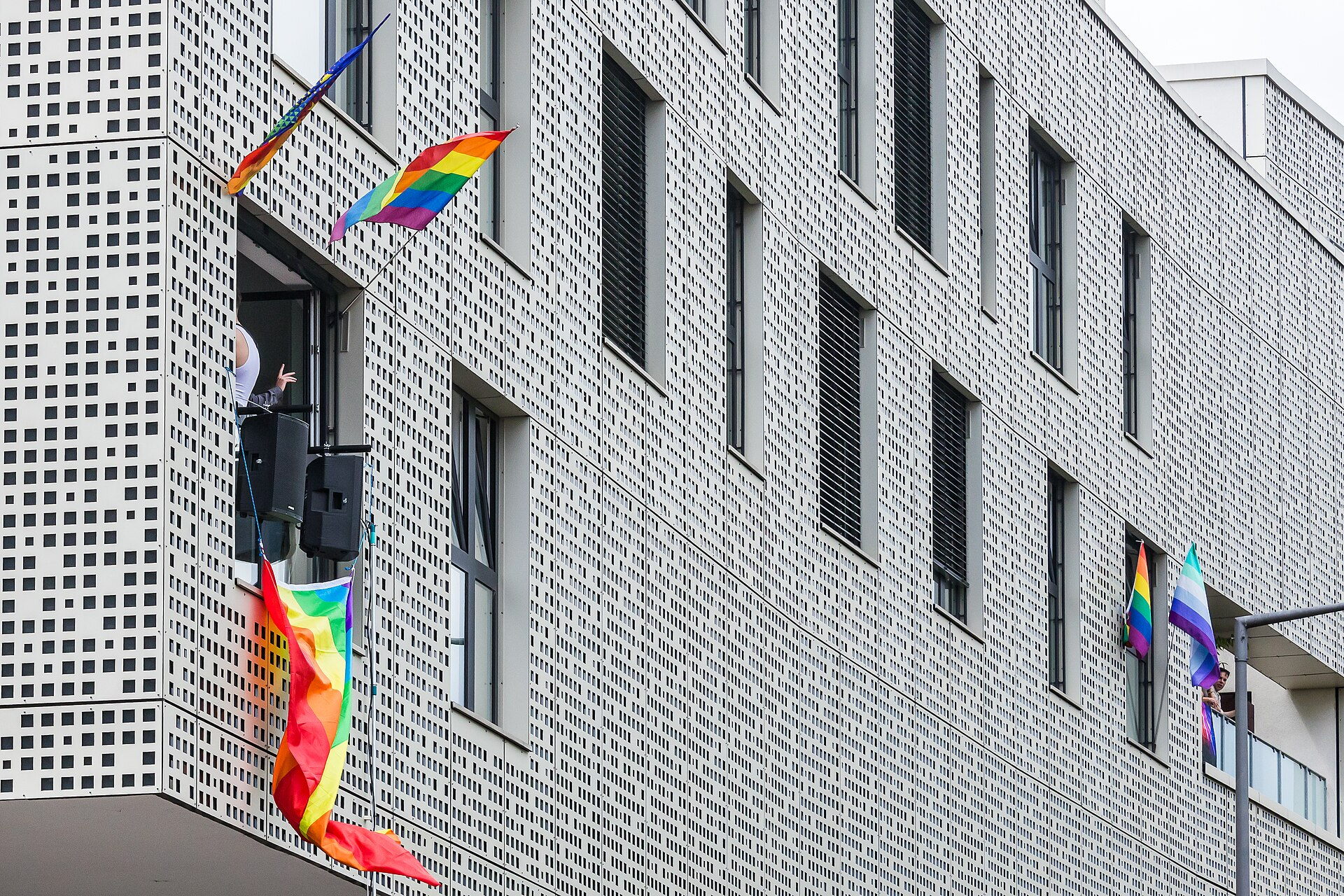 Links: ein offenes Wohnungsfenster. Darin stehen zwei große Lautsprecher und es hängen Regenbogenfahnen aus dem Fenster