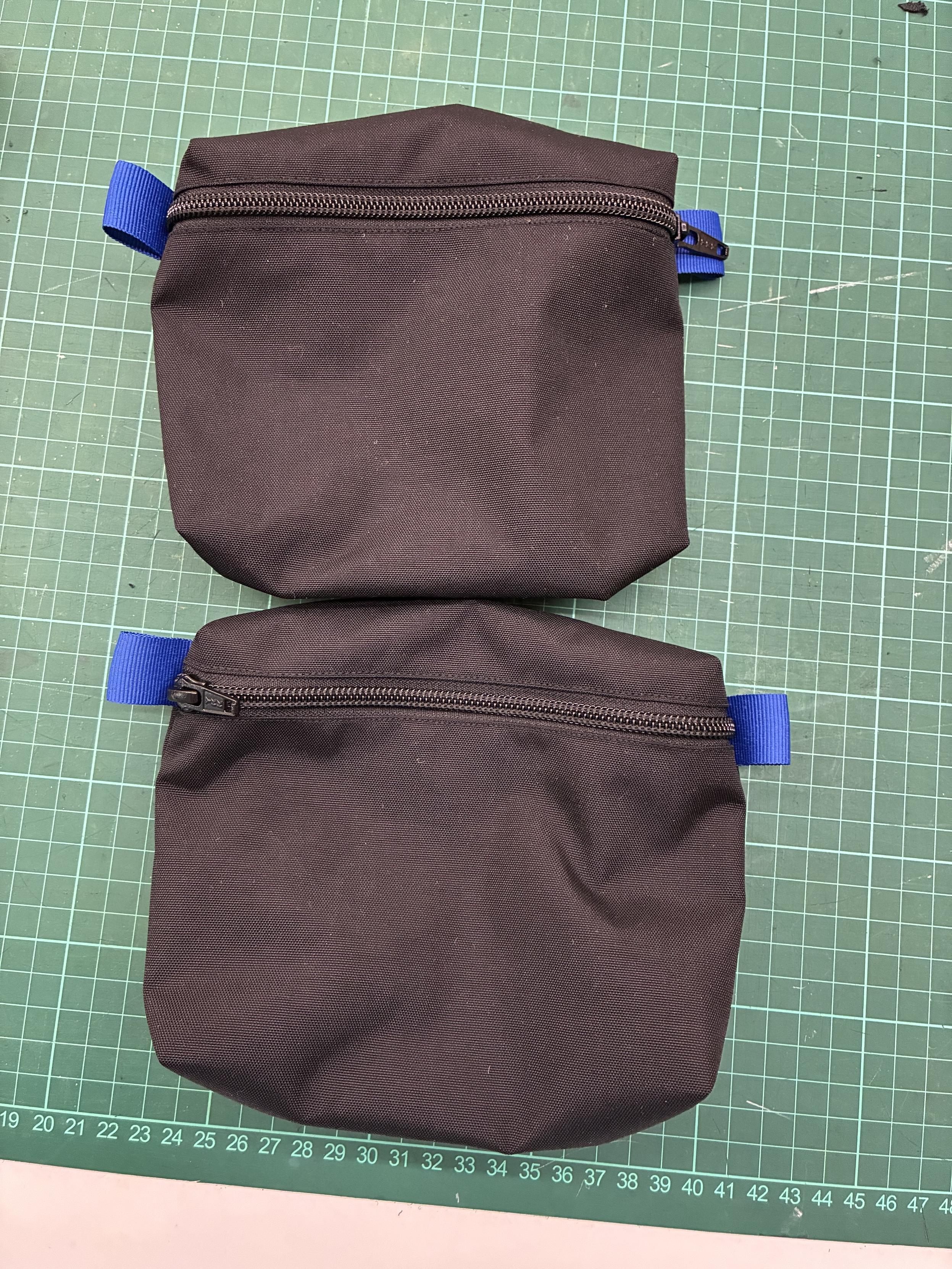 Two black fabric pouches with blue zipper pulls, placed on a green cutting mat with grid lines.