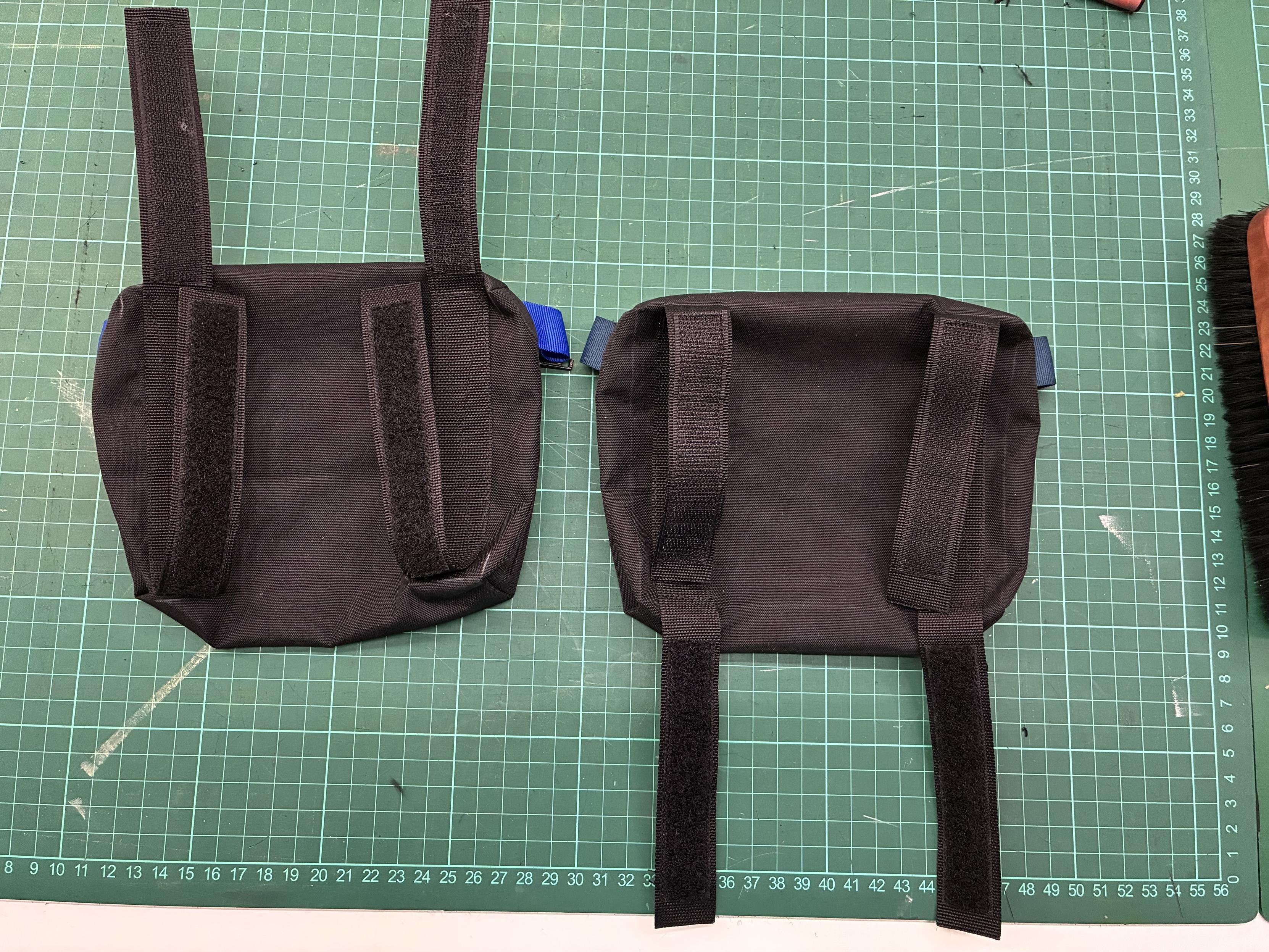 Two small black fabric bags with straps, placed on a green cutting mat. The bags feature Velcro on the sides and are designed for attachment or carrying purposes.