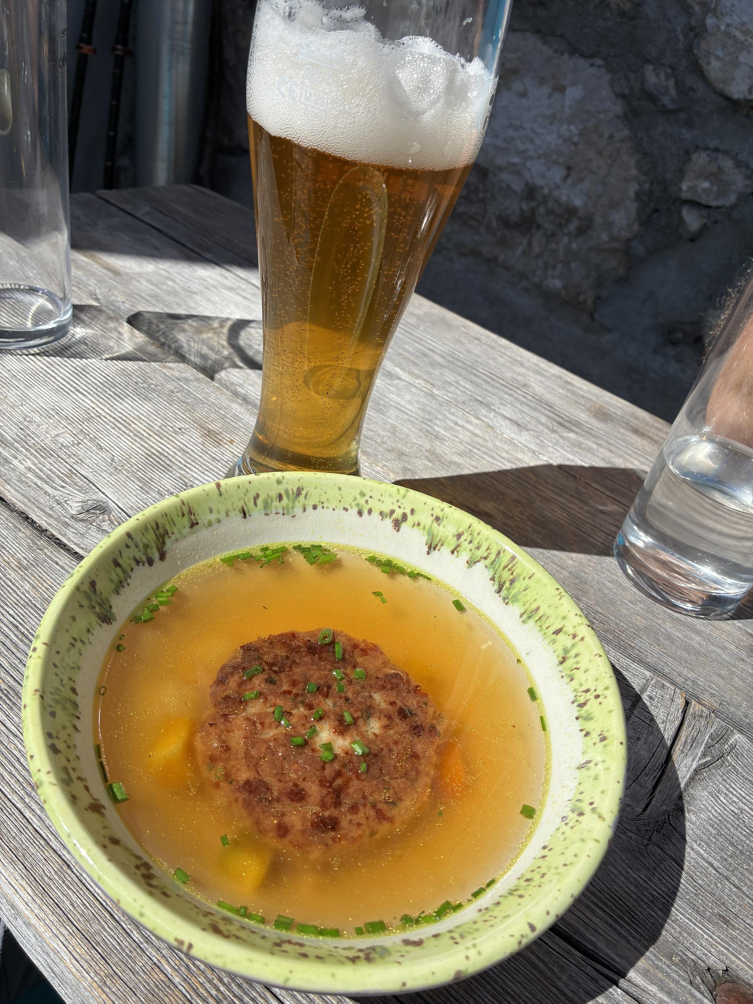 A bowl of soup containing a kaspressknödel, garnished with chopped chives, is placed on a wooden table. Next to it, a tall glass of golden beer with a frothy top is visible.