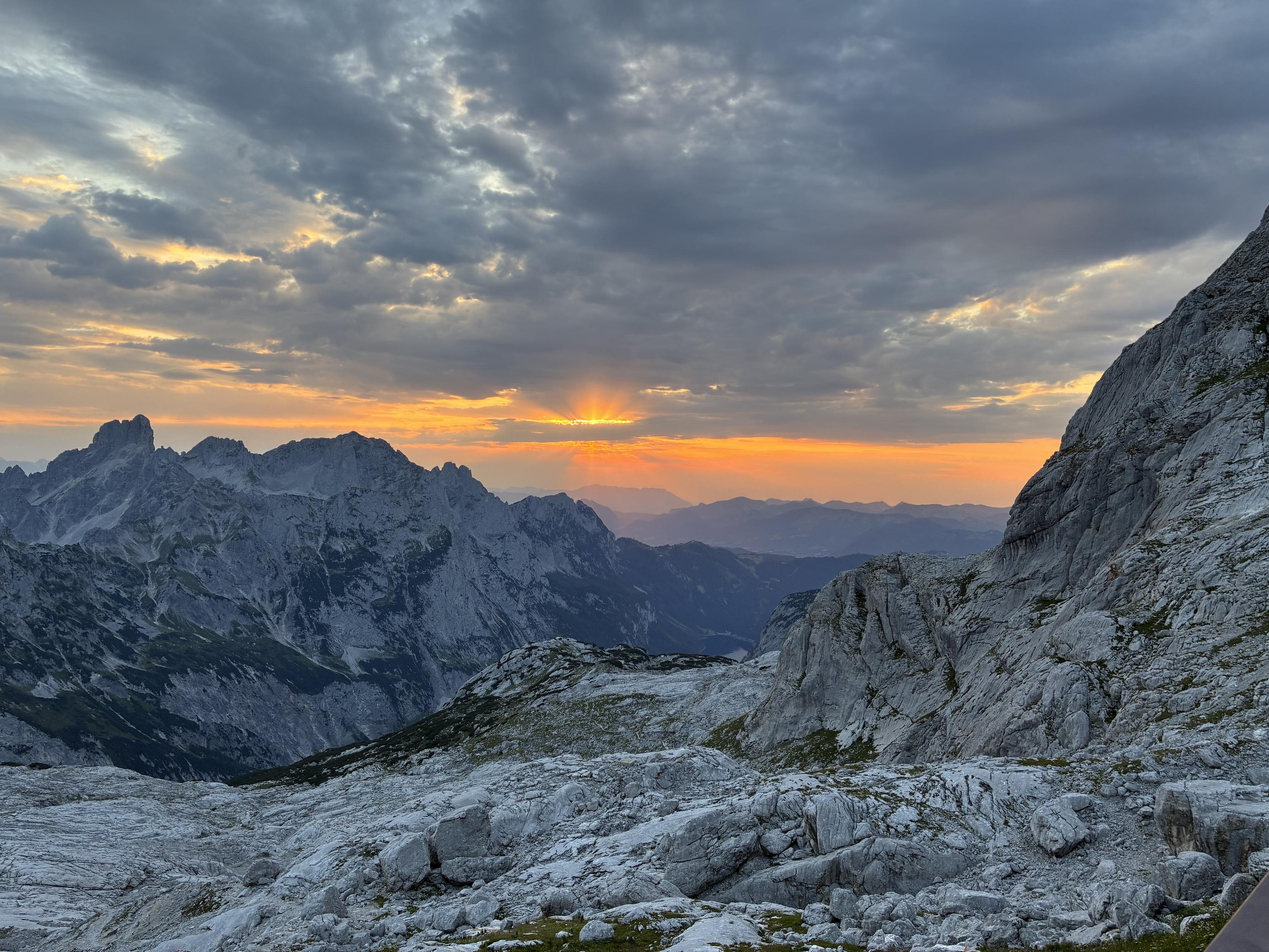 A breathtaking mountainous landscape at sunset, featuring rugged peaks silhouetted against a colorful sky. The foreground showcases rocky terrain and patches of greenery, while the sun sets behind the mountains, casting a warm glow.