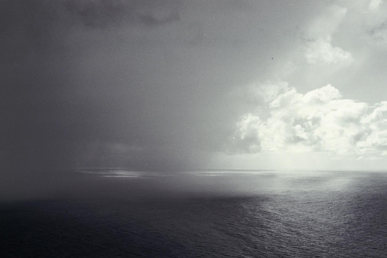 das schwarzweißbild zeigt eine weite ansicht des atlantischen ozeans unter einem dramatischen himmel.
die linke seite ist von dunklen, schweren regenwolken dominiert. ein sichtbarer regenschauer zieht dort dicht über das wasser. auf der rechten seite bricht helles sonnenlicht durch die wolkendecke. helle, kontrastreiche wolkenformationen heben sich dort ab. das sonnenlicht spiegelt sich in hellen streifen auf der meeresoberfläche. das wasser hat eine unruhige struktur mit feinen wellen.