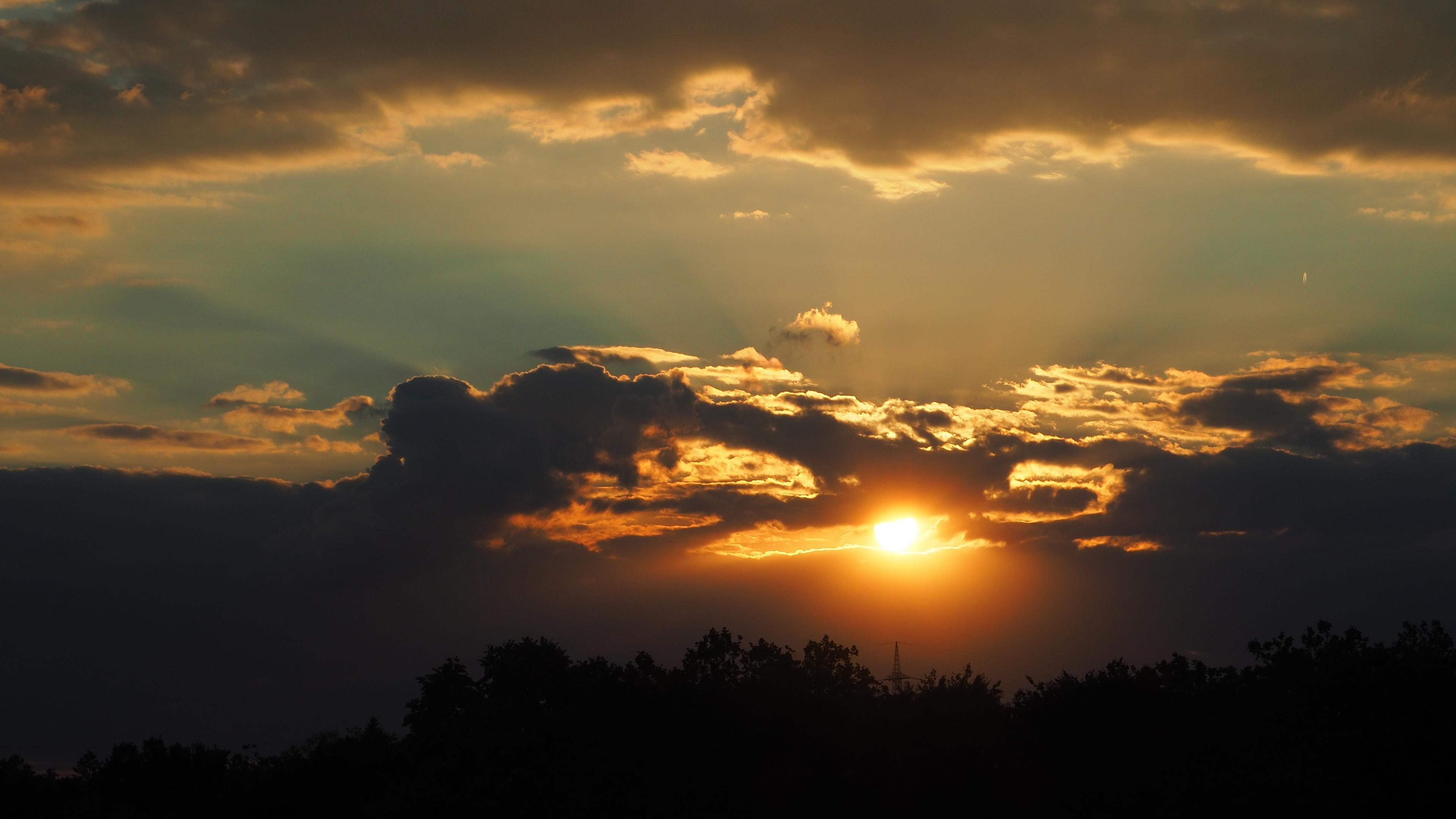 Sonnenuntergang hinter Abendwolken.
Am unteren Bildrand eine fast schwarze Baumreihe.
Darüber eine Wolkenbank mit Lücken. Durch eine scheint die fast untergegangene Sonne.
Daüber ein schmales Band unbewölktem fahlblauen Himmel und darüber wieder Wolken.