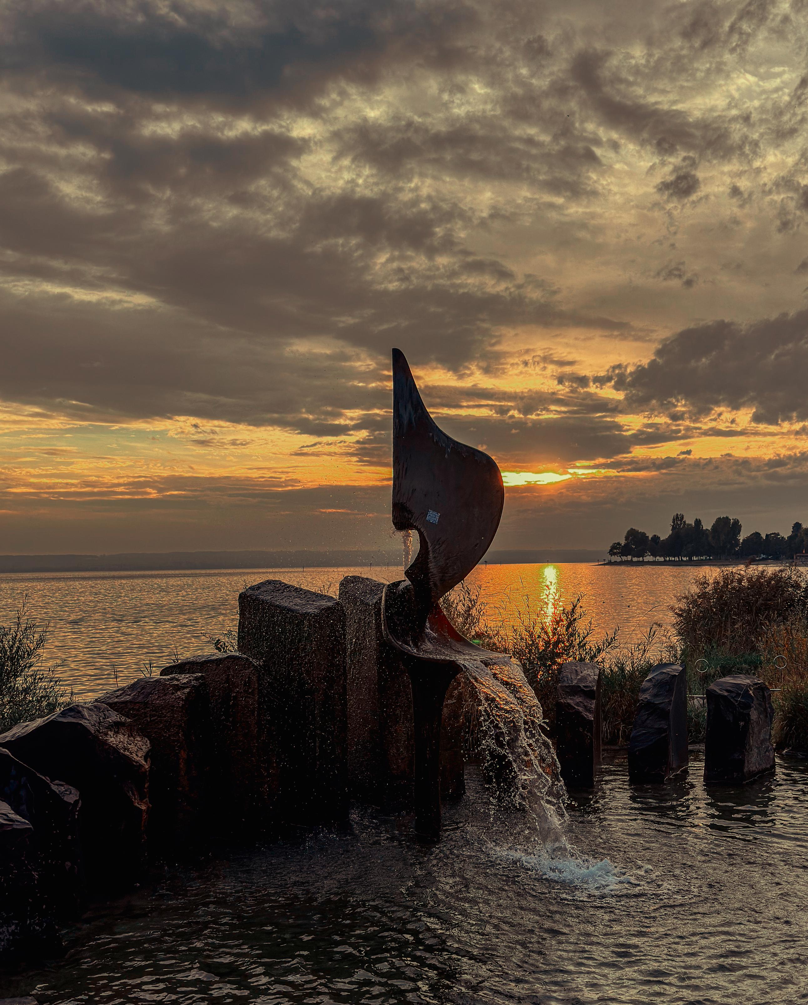 Abendstimmung am See. Sonnenuntergang in warmen Farben. In der Mitte eine Skulptur als Springbrunnen.