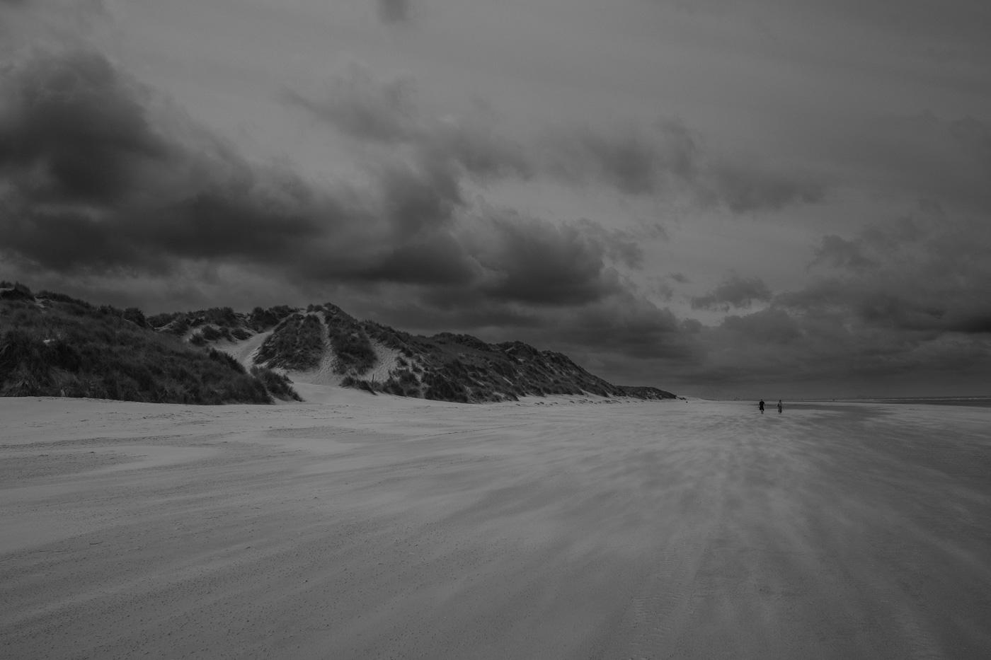 Starker Wind bläst den Sand über den breiten Strand, rechts sind 2 Spaziergänger zu sehen. Links Sanddünen die mit Gräsern bewachsen sind. Der Himmel ist sehr stark bewölkt. Foto ist in schwarz-weiß