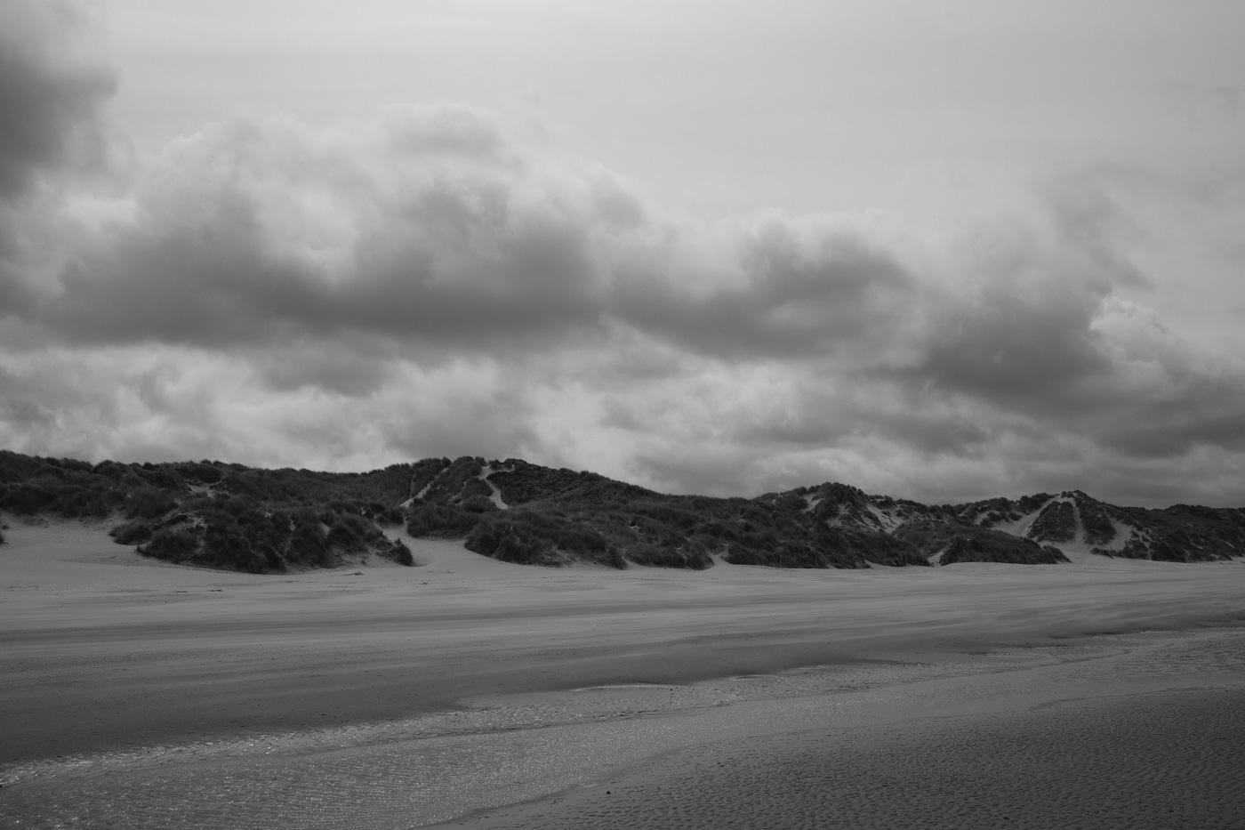 Ein breiter Sandstrand mit Dünen im Hintergrund, der Himmel ist stark bewölkt. Schwarz-weiß Foto.