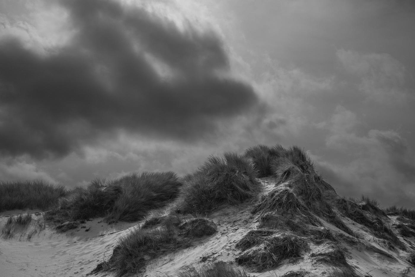 Die Spitze einer Sanddüne mit den typischen Gräsern bewachsen. Der Himmel ist stark bewölkt, mit dunklen Wolken und kleineren Lücken. Foto ist in schwarz-weiß.