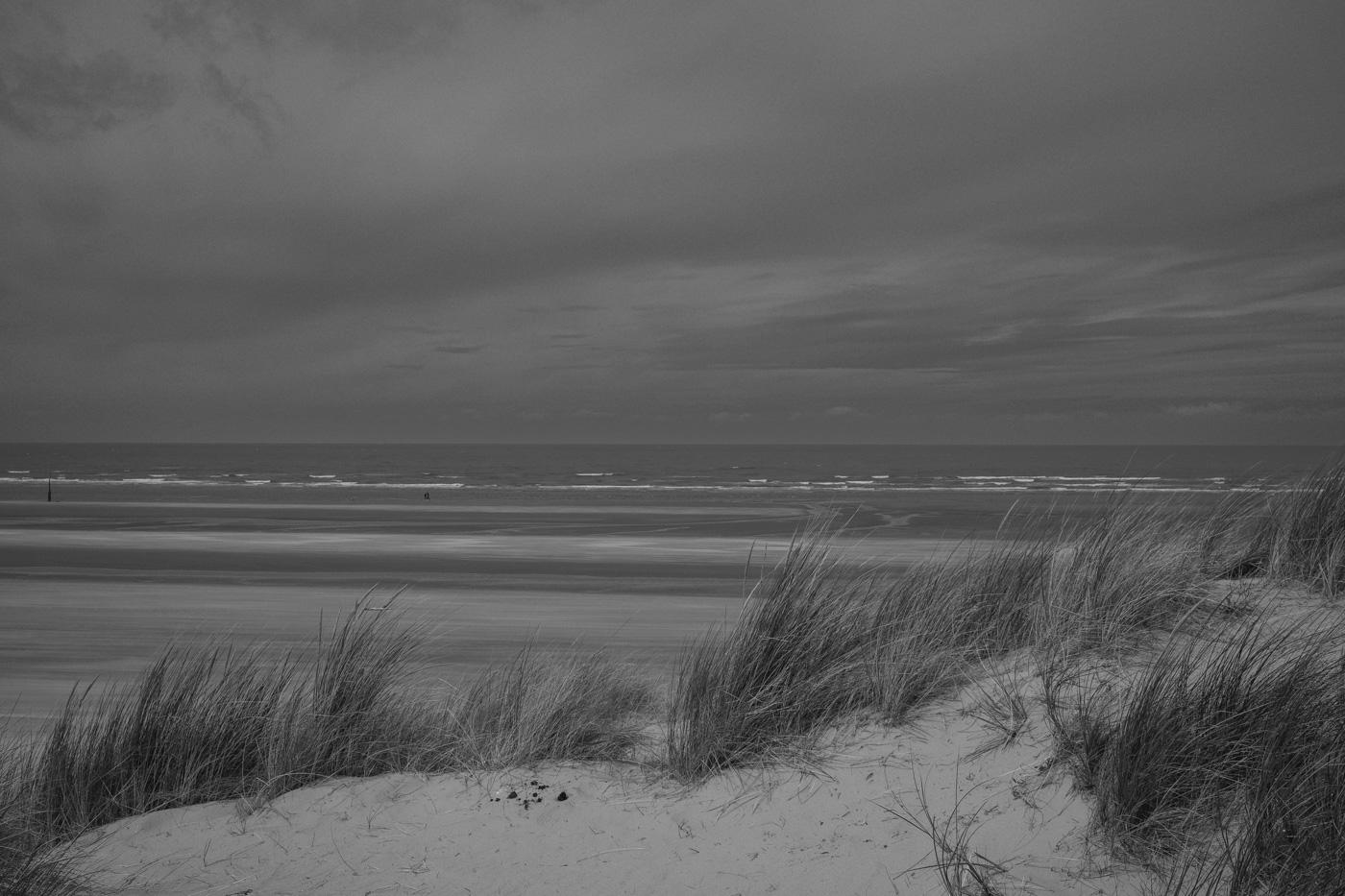 Ein Foto von einer Düne auf das Meer hinaus, das mit leichten Wellen an den Strand gespült wird. Der Himmel ist stark bewölkt. Foto in schwarz-weiß.