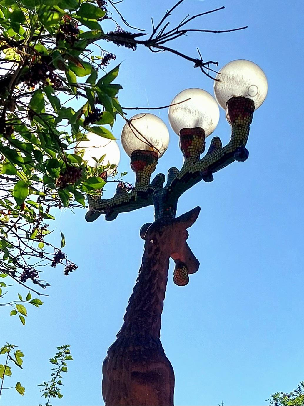 A half-rear view of a large street lamp. It is shaped like a stag and decorated with small, colourful mosaic tiles. Its four-pointed antlers are crowned by four glass balls. The light bulbs inside cast a soft glow into the blue twilight. A couple of green twigs stretch into the picture from the left. A dreamy, almost fairy-tale atmosphere.