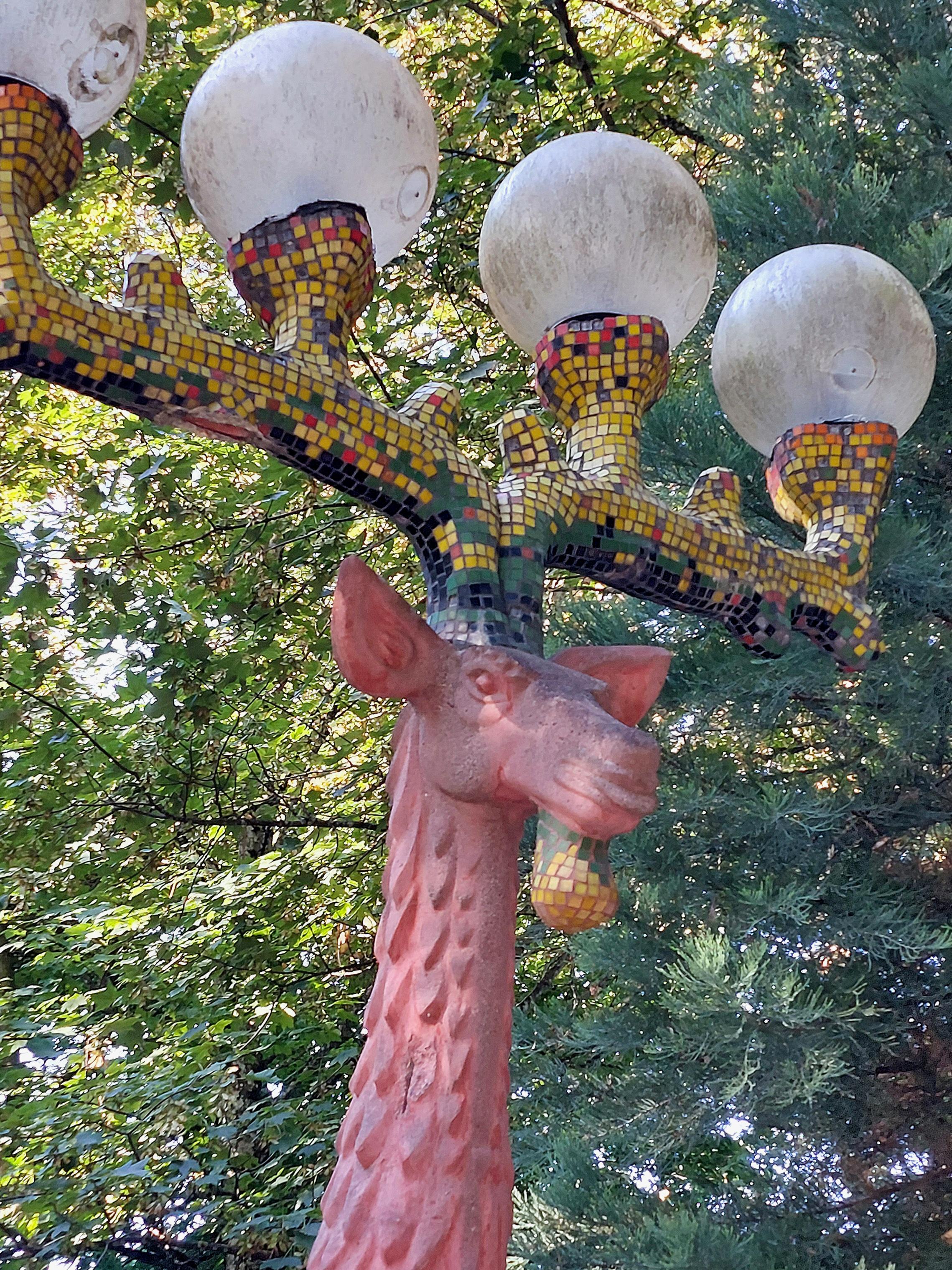 StraßenLaterne in Form eines Hirsches. Auf dem Geweih sitzen vier Glaskugeln, welche die Leuchtmittel enthalten. Material: roter Sandstein mit bunter Mosaikverzierung. Grüne Bäume im Hintergrund. Helles Morgenlicht. 
  //  A street lamp in the shape of a rearing stag. Four glass globes, containing the light sources, sit on the antlers. Material: red sandstone with colourful mosaic decoration. Green trees in the background. Bright morning light.