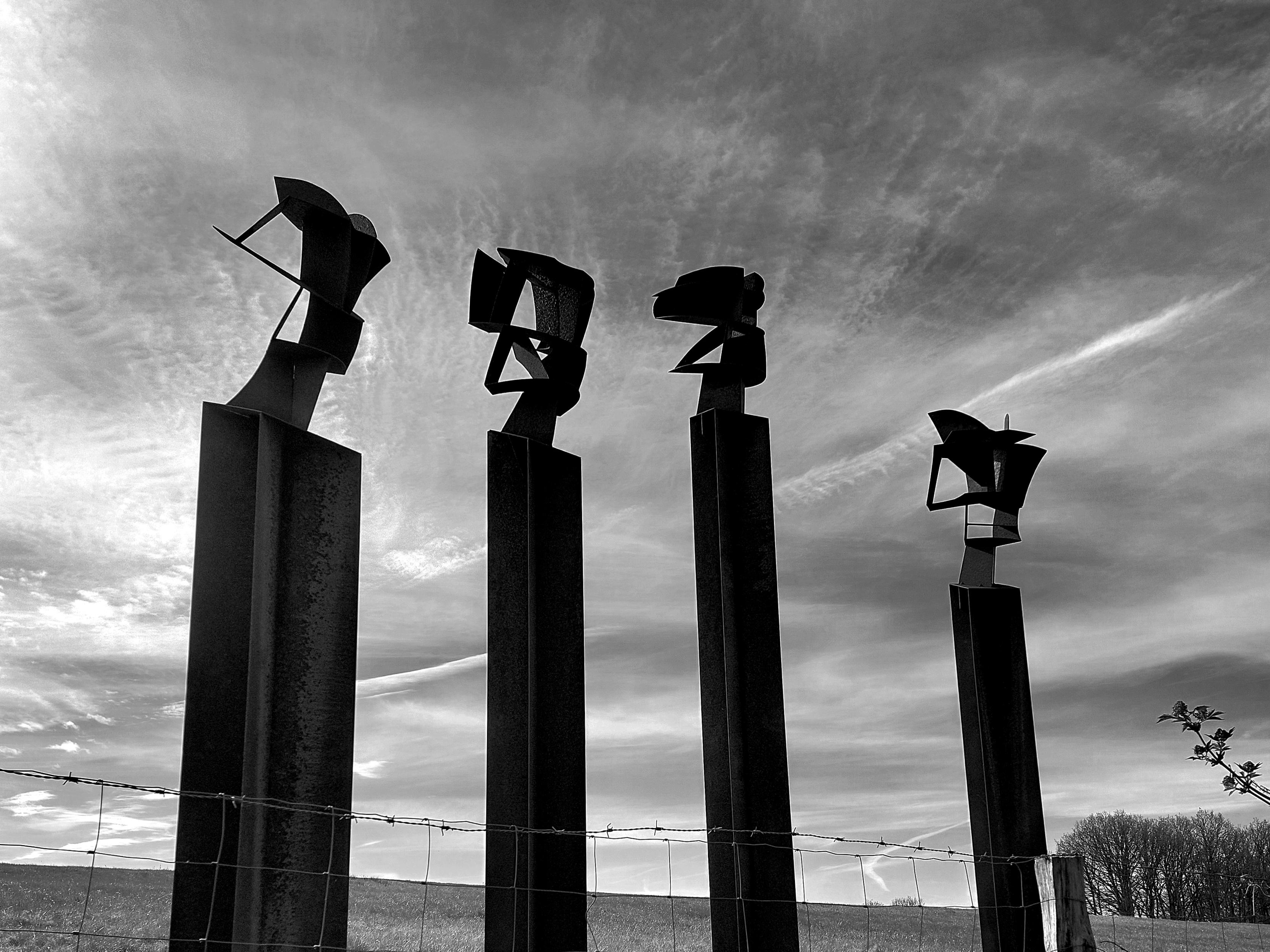 Black-and-white shot, landscape format. Against a dramatic sky with wispy clouds and con trails four dark sculptures stand on tall, slender columns. They embody the inner demons, fears and hidden shadows that everyone carries within themselves.